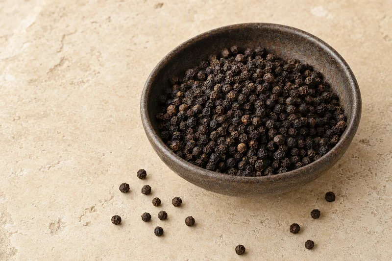 Whole black pepper in a ceramic bowl; illustrative product image